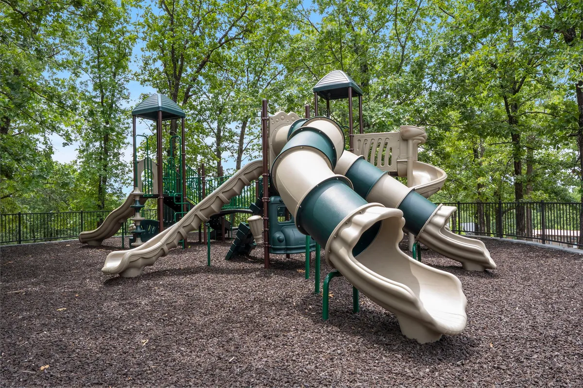 A vibrant playground featuring slides and a climbing structure surrounded by tall trees. Children are playing on the equipment, and families are gathered nearby, creating a lively and joyful atmosphere.