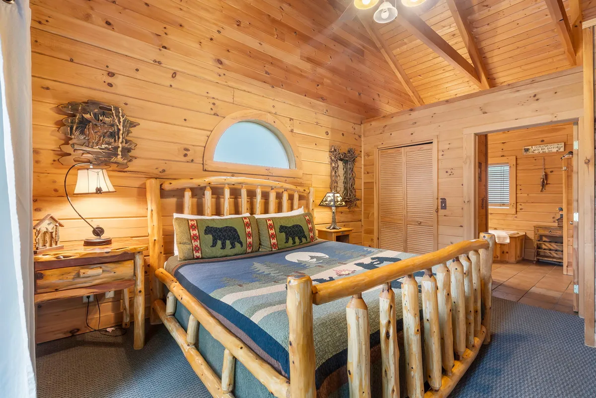A warm, rustic master bedroom featuring a log bed adorned with plush, bear-themed pillows, surrounded by handcrafted wooden furniture and decorative wall art. Soft lighting and cozy blankets invite relaxation, while views of the outdoors add to the tranquil ambiance.