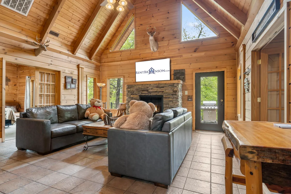 This image showcases a spacious living area with large windows letting in abundant natural light, a substantial stone fireplace, plush teddy bears on the couch, wooden accents, and a flat-screen TV. The room exudes a warm, family-friendly atmosphere perfect for relaxation and group activities.