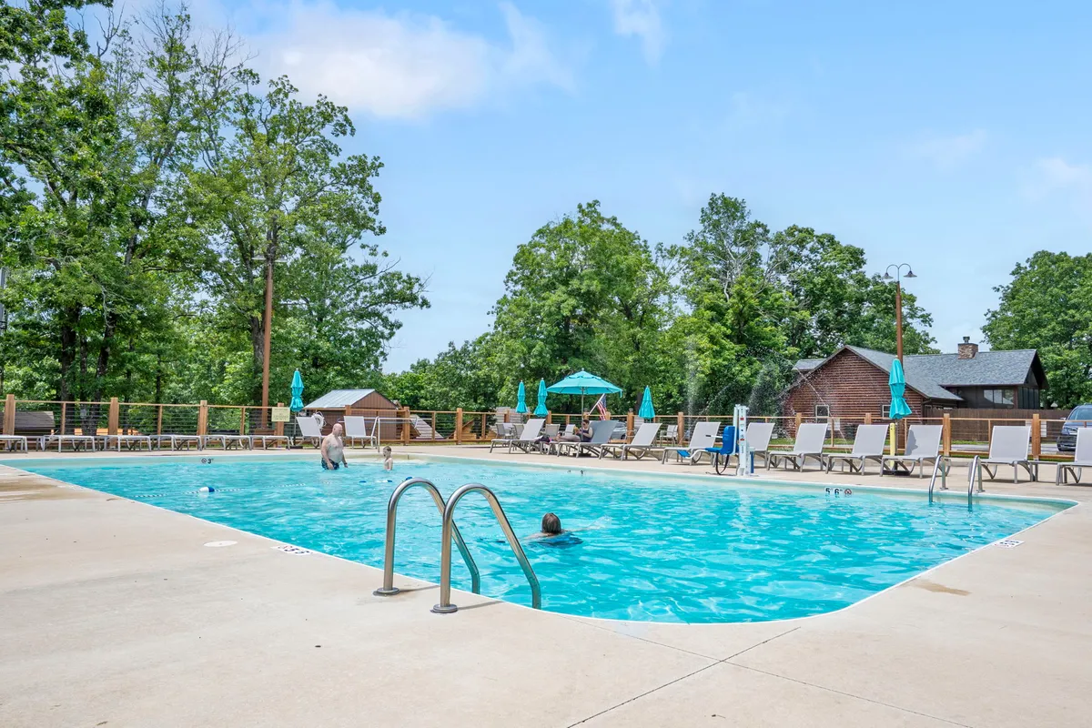 A bustling resort swimming pool framed by lounge chairs, umbrellas, and lush greenery. People are swimming and relaxing poolside, while the sun casts a warm glow across the water and trees.