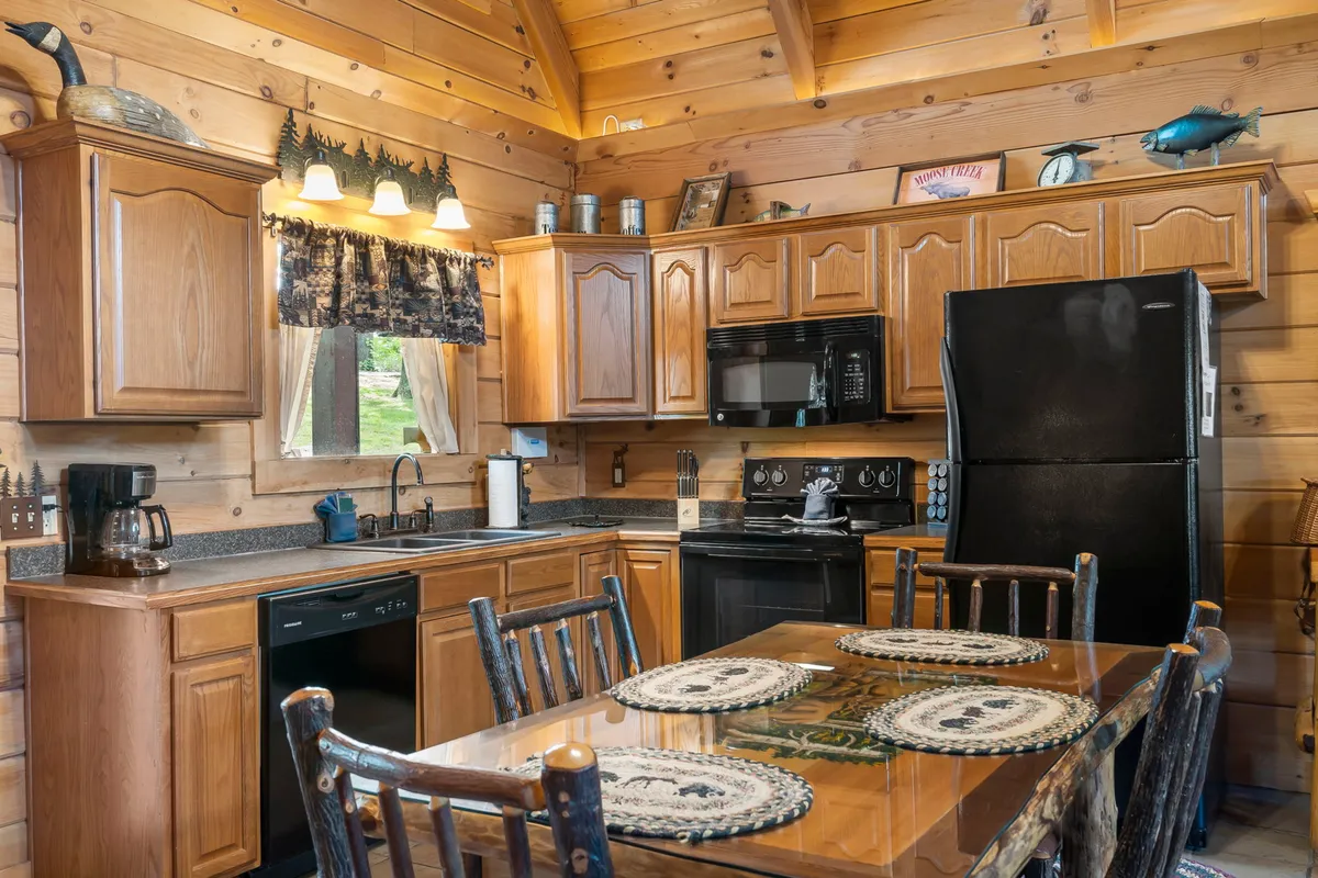 This image showcases a warm and inviting kitchen and dining space, featuring a kitchen island seamlessly integrated with a spacious dining table. The area is enhanced by wooden cabinetry, black modern appliances, and decorative accents like fish and quirky signs. The setting is ideal for both preparing meals and gathering with friends or family for group activities.