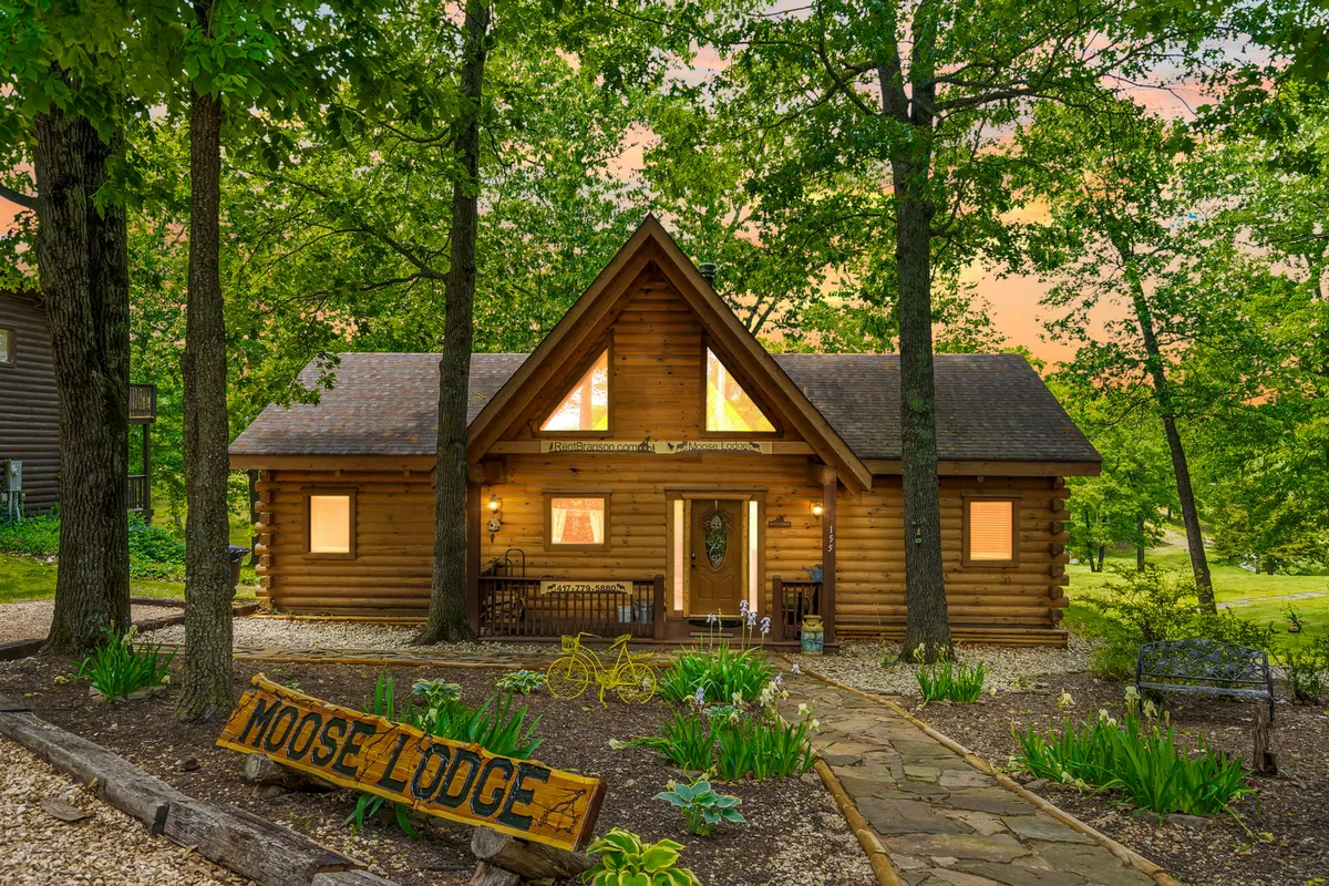 The hero image features a charming log cabin with a welcoming 'Moose Lodge' sign, nestled amidst a beautifully landscaped garden. A stone pathway leads to the front porch, perfect for relaxing and enjoying the peaceful natural surroundings. The cozy exterior invites guests to imagine outdoor gatherings, sunset views, and tranquil moments in nature.
