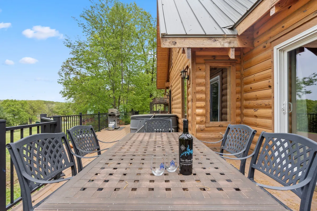 A welcoming outdoor dining area set with a bottle of wine and glasses atop a wooden table, nestled among lush green trees. The scene radiates a sense of privacy and intimacy, perfect for couples or small groups savoring alfresco meals together. The setting is relaxed, with sunlight dappling through the leaves and a gentle breeze hinted at by the movement of the foliage.