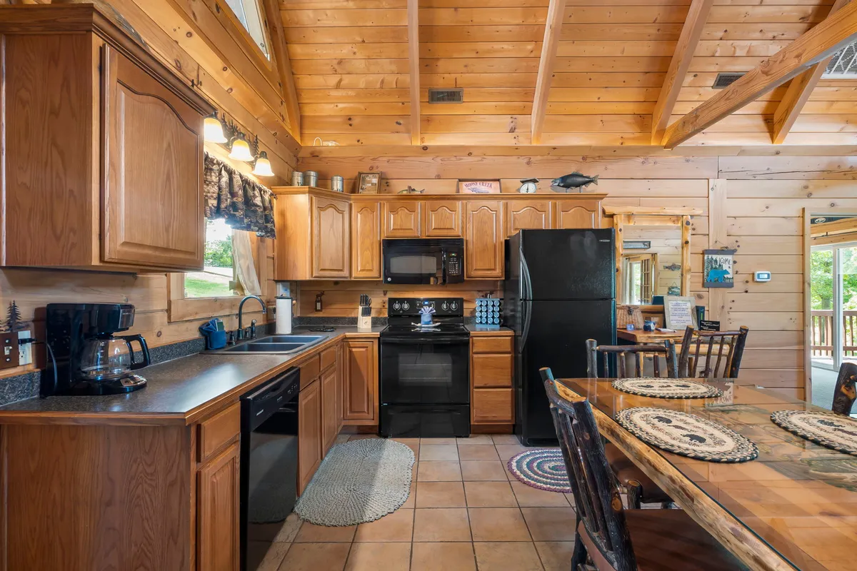 This interior image highlights a cozy kitchen and dining environment, with wooden cabinets, black appliances, a roomy dining table, and large windows that let in natural light. The space is set up to accommodate family gatherings, cooking sessions, and memorable meals.