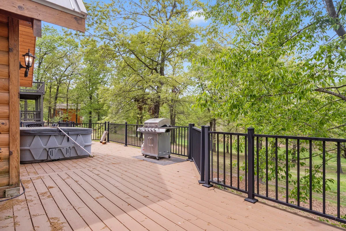 A spacious wooden deck set amidst lush trees, featuring a prominent hot tub, a propane grill, and an outdoor dining area. The atmosphere is inviting and relaxed, perfect for unwinding or hosting outdoor meals. The hot tub is a centerpiece, suggesting cozy evenings under the stars, while the grill and dining table promise al fresco feasts.